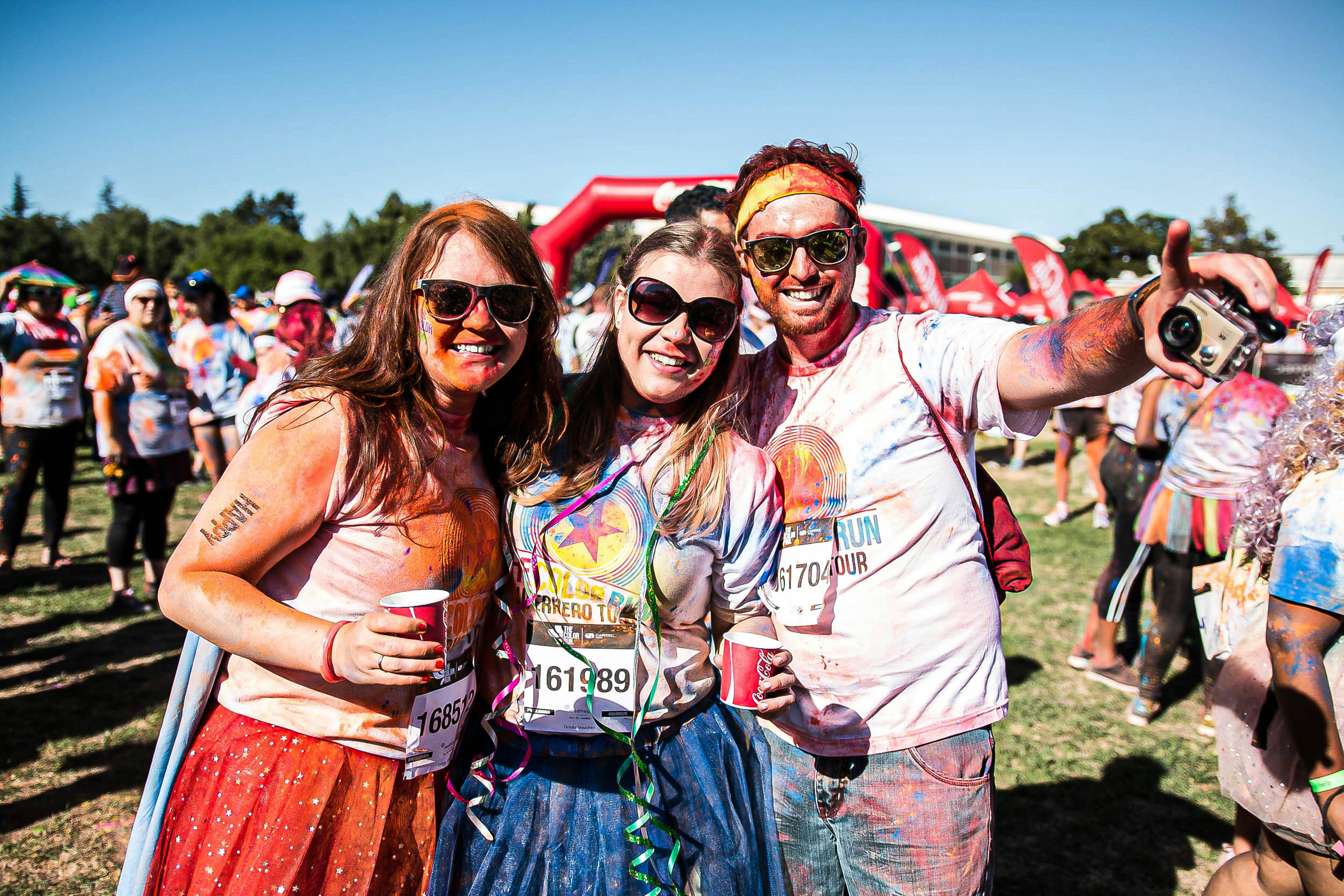 Trio of people in costume props and covered in brightly coloured powder smiling at the camera. Other event participants and the inflatable finish line can be seen in the background.