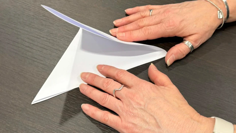 The image shows a person folding a triangle made from paper into a smaller triangle. One hand is holding the paper steady on the table, while the other hand is folding one corner over to create the new shape.
