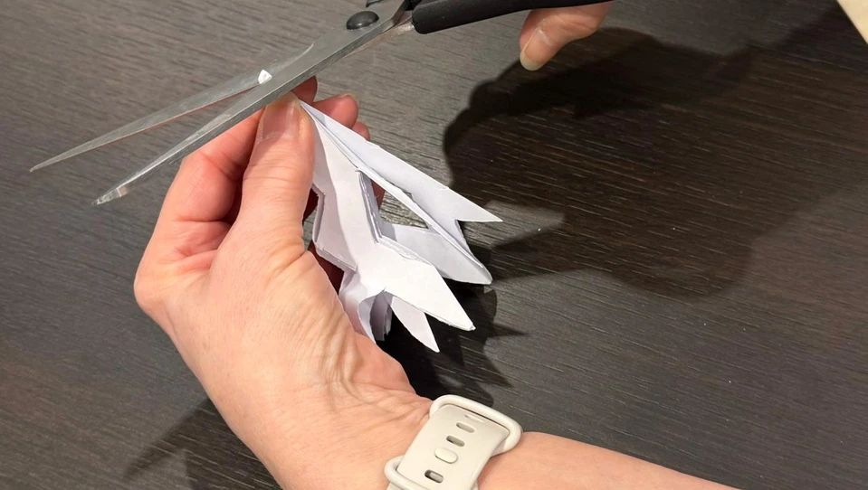 The image shows a person using scissors to carefully cut shapes into the folded paper snowflake. Their other hand holds the folded triangle steady. The cuts are decorative, creating the intricate patterns that will appear when the snowflake is unfolded.
