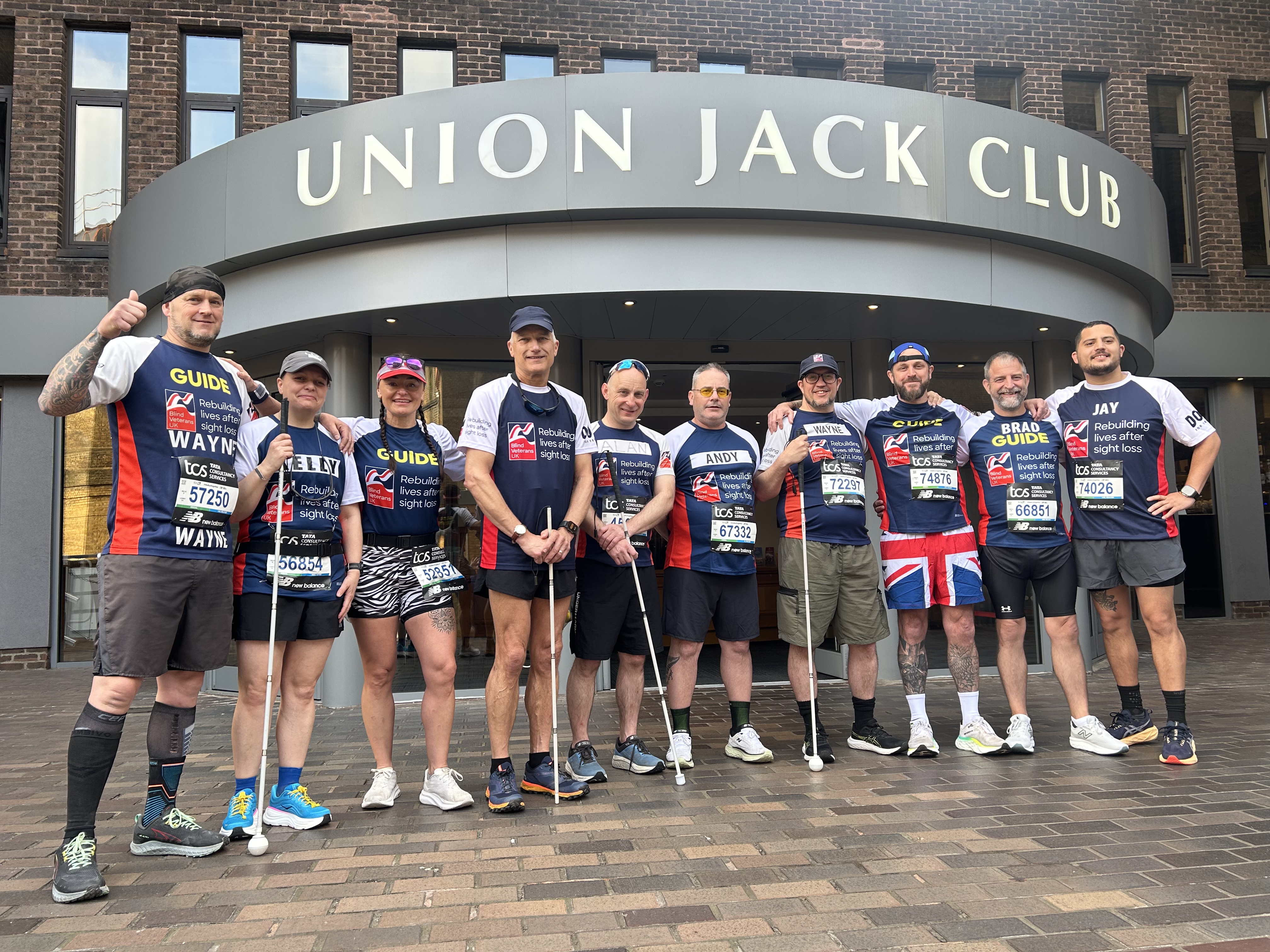 A group photo of our veterans and their guides outside the front of the Union Jack club on the morning of the London Marathon. The group are all in their running gear and displaying their running numbers.