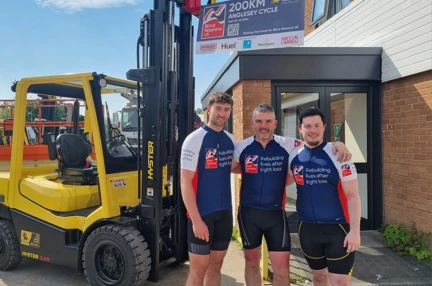The three riders in their cycling tops stood in front of a fork lift truck at their workplace and a banner with the Blind Veterans UK logo and 200km Anglesey Cycle written on it