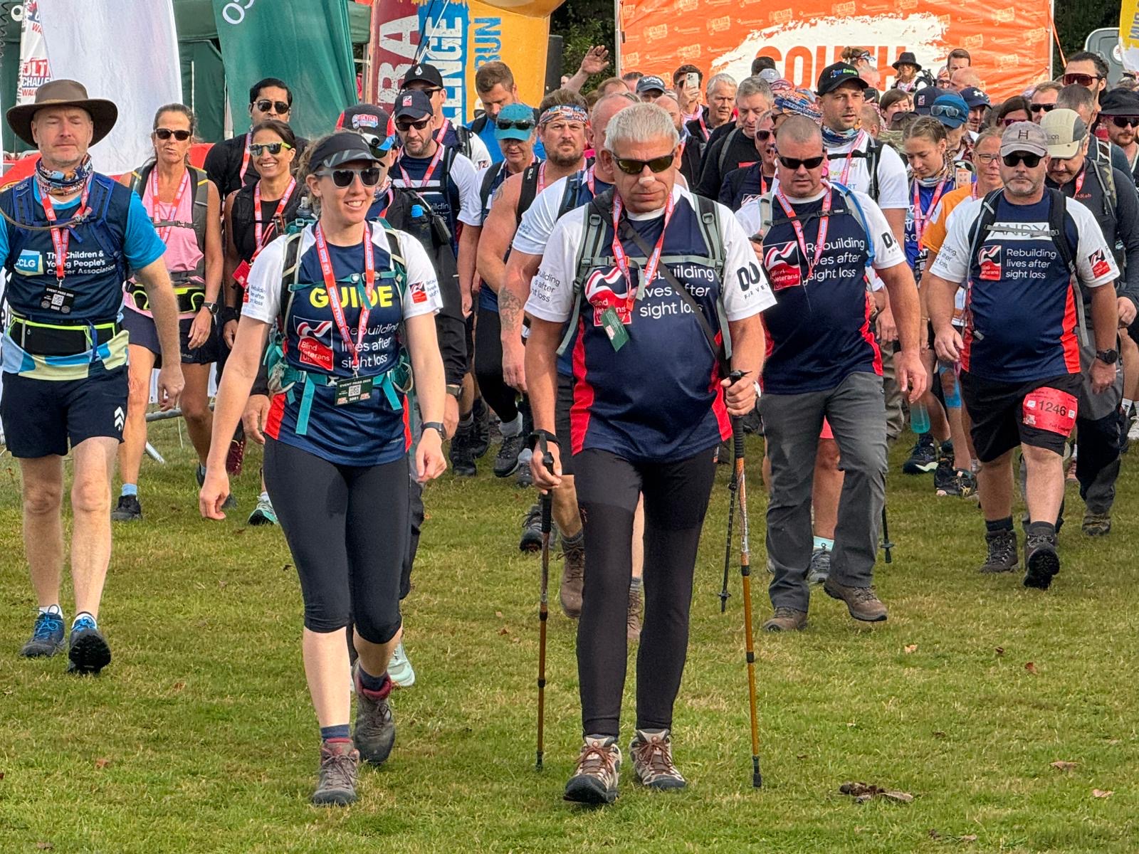 A team of blind veterans and guides wearing their Blind Veterans UK tops confidently striding away from the start line with a crowd of participants following them.
