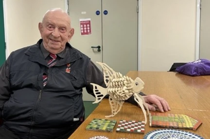 Glynne sat next to his creative projects – A wooden model of an angelfish, surrounded by colourful mosaics
