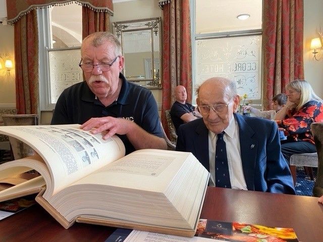 Matt is sitting at a table as a man next to him turns the page on a giant book