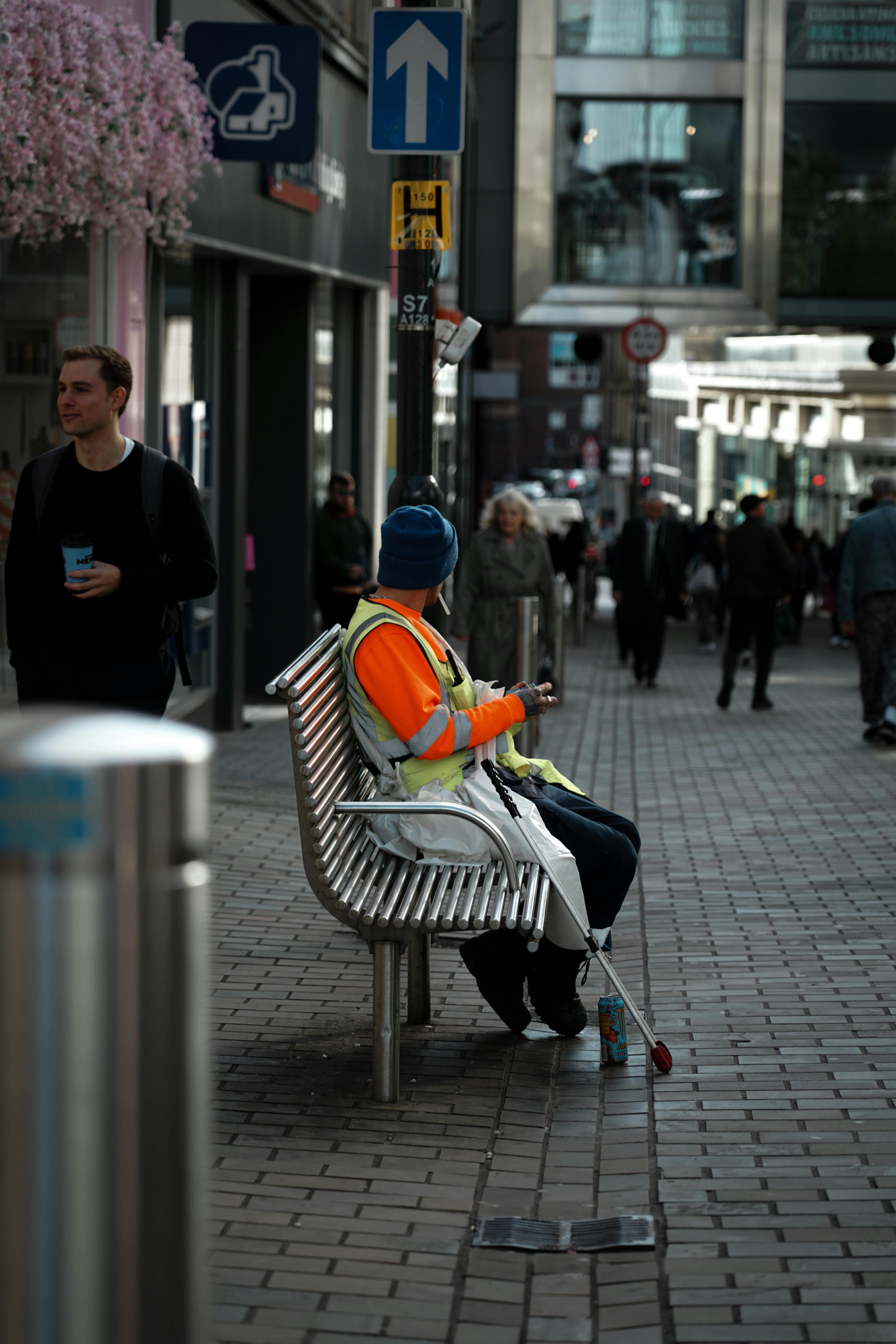 A vision-impaired person sitting on a high street bench, wearing a high-visibility vest and holding a white cane