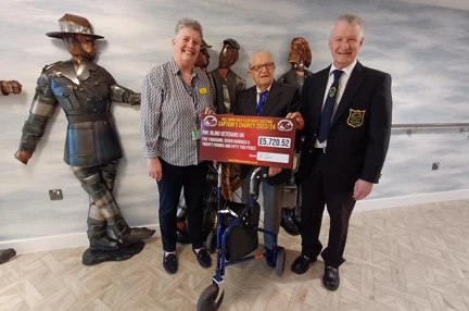 Blind veteran Les is stood between Lesley and his son Keven as they present Lesley with a large cheque