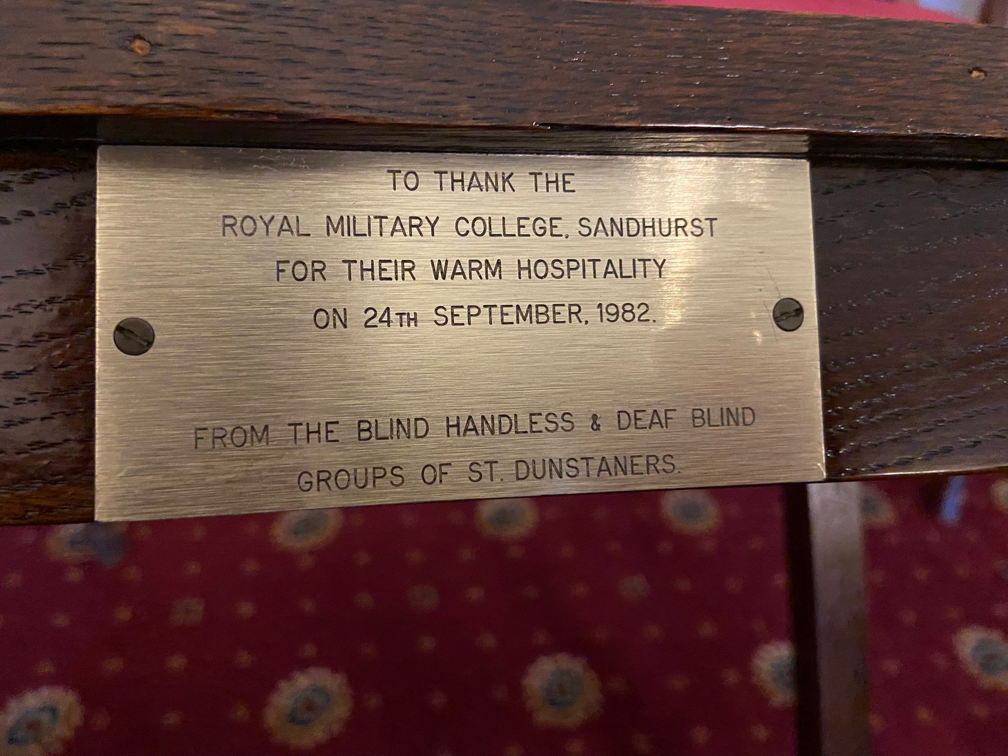 Close-up of a brass plaque fixed to a wooden table, engraved with thanks to the Royal Military College Sandhurst for warm hospitality on 24 September 1982 from St Dunstaners.