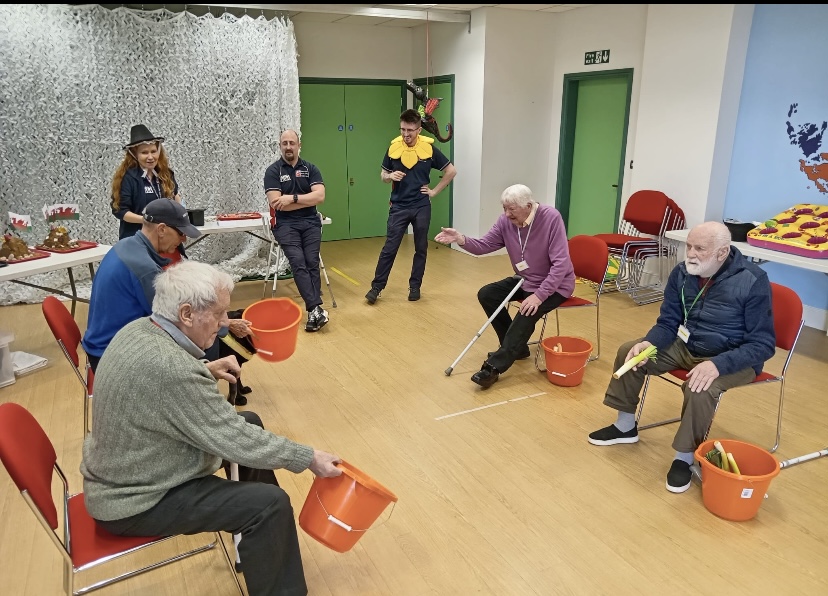 A group of people sit on chairs in a circle holding buckets and attempt to throw leeks into the buckets