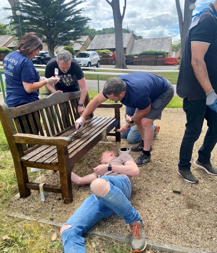 Four Thea colleagues working together to varnish a wooden garden bench at our Blind Veterans UK Llandudno Centre of Wellbeing. One of the group is lay on the floor so he can reach underneath.