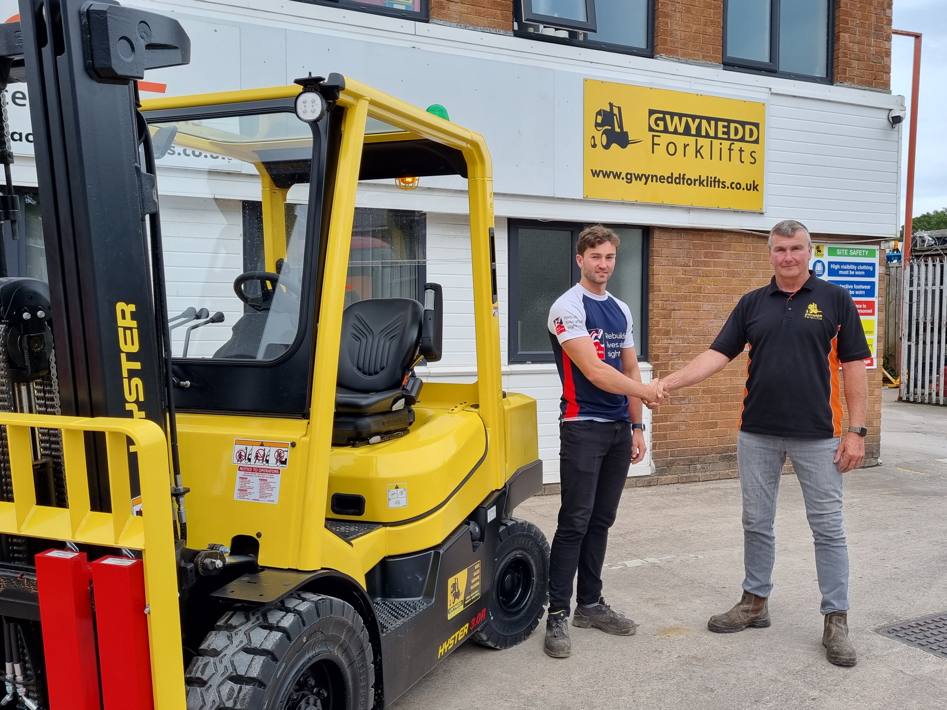 James stood in front of a forklift truck wearing his Blind Veterans UK t shirt shaking hands with his dad who is director of Gwynedd Forklifts Ltd