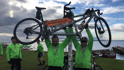 A photo of Craig standing outside holding a tandem bike overhead with his teammate