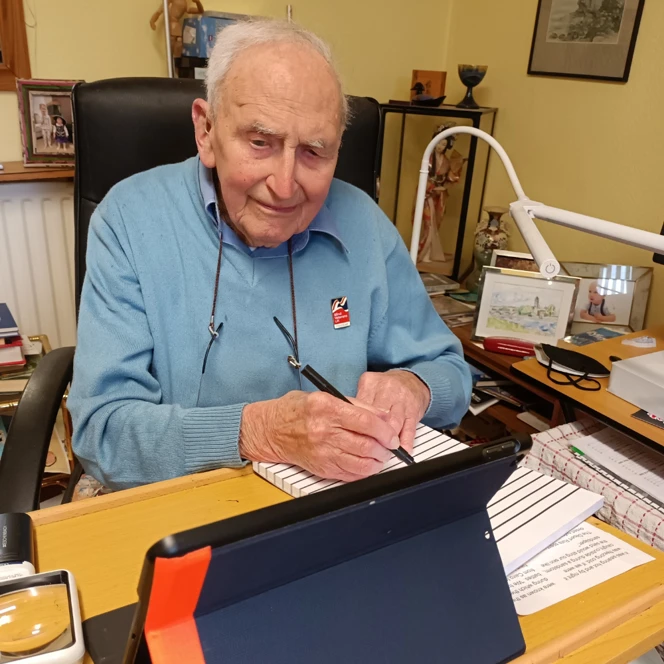 John is sat at a desk holding a pen and writing on wide lined paper