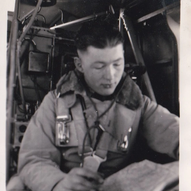 A black and white photograph of John, seated inside an aircraft. He appears focused, looking down, possibly reading or looking at a map.