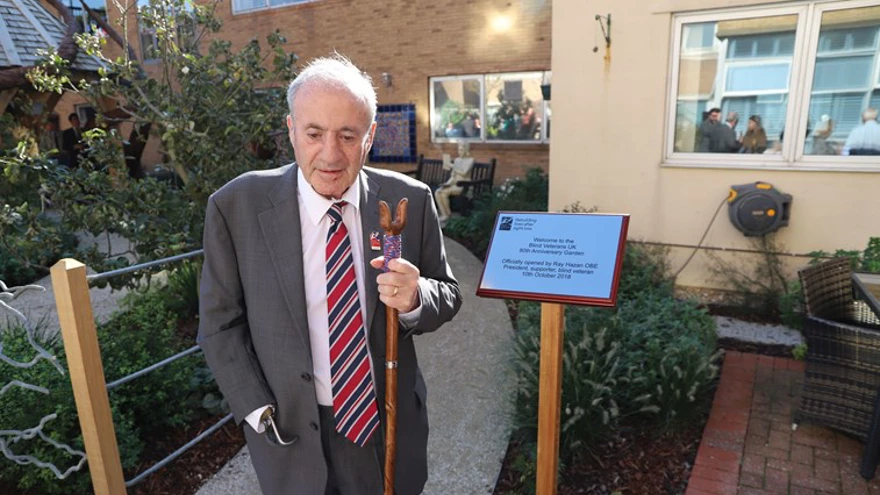 Blind veteran Ray Hazan OBE, holding a walking stick, standing in the garden of our centre of wellbeing, next to a plaque commemorating the opening of the garden.