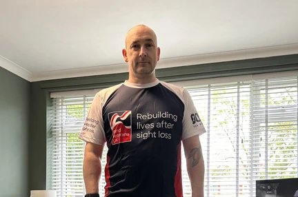 Blind veteran Steve standing in his living room wearing his Blind Veterans UK t-shirt